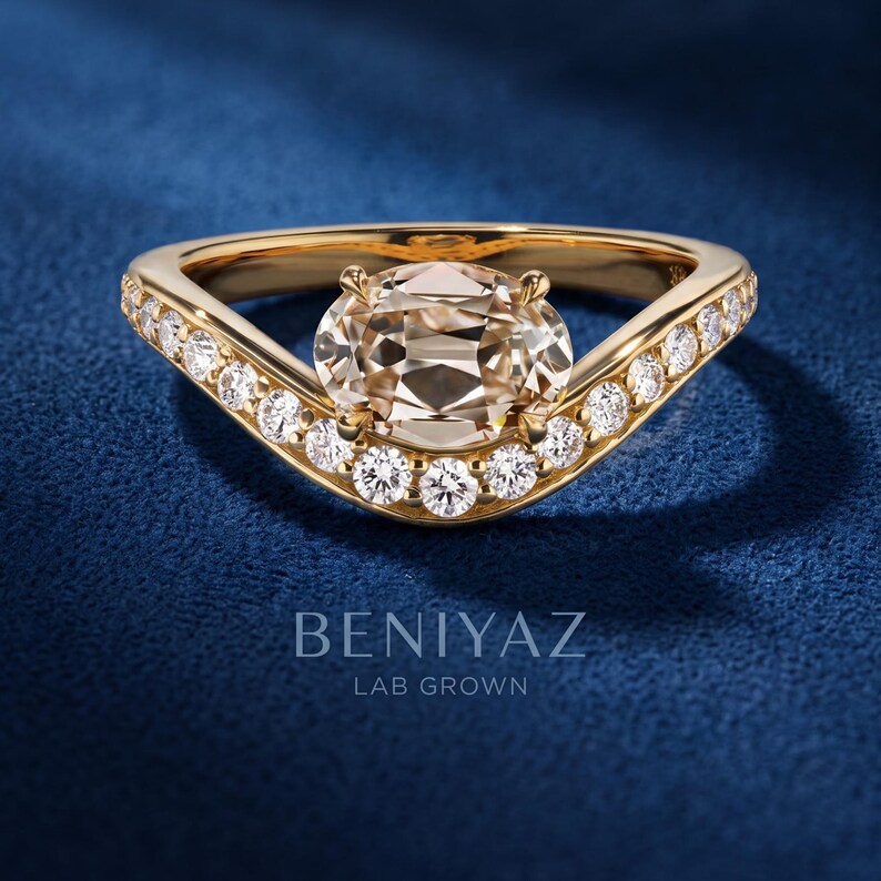 Macro side-angle photo of an oval cut lab grown diamond ring held between fingers, highlighting the yellow gold setting, center stone and pavé diamond bypass band.