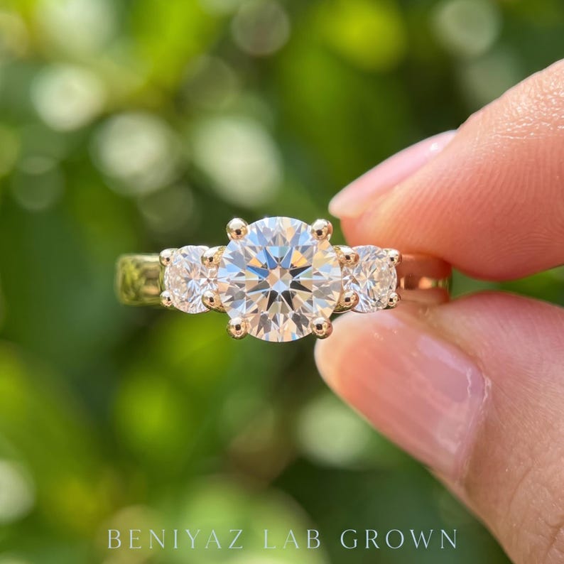 May include: A three-stone ring featuring a large round brilliant-cut centre diamond flanked by two smaller round diamonds, all set in a polished gold band. The text BENIYAZ LAB GROWN is printed at the bottom of the image.