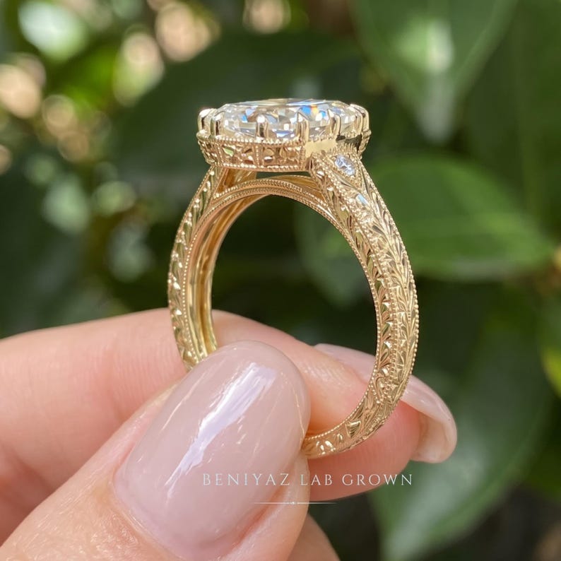 Side profile of an oval diamond engagement ring held upright between fingertips. This view shows the elevated center stone setting, decorative shoulders and diamond-accented band against green foliage.
