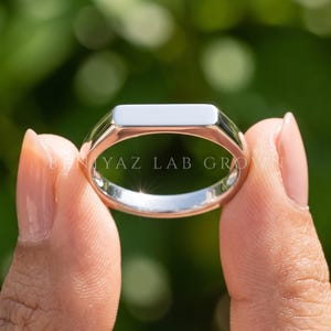 May include: A polished silver-toned signet ring featuring a flat, rectangular top surface. The band is smooth and reflective, showcasing a clean, minimalist design. The text BENIYAZ LAB GROWN is clearly visible across the center of the ring.