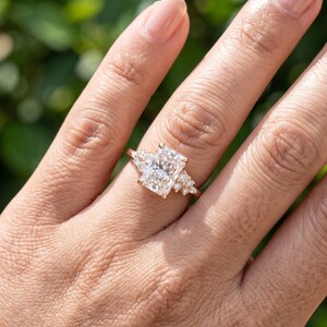 May include: A rose gold ring featuring a large rectangular radiant-cut center stone flanked by a cluster of three smaller round-cut stones on each side. The band is thin and polished, showcasing a clear, faceted design that catches the light.
