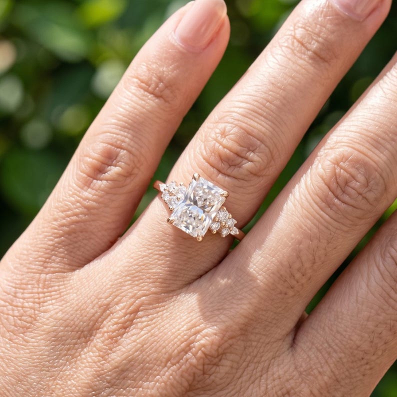 May include: A rose gold ring featuring a large rectangular radiant-cut center stone flanked by a cluster of three smaller round-cut stones on each side. The band is thin and polished, showcasing a clear, faceted design that catches the light.