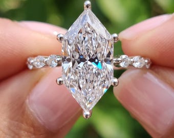 Puede incluir: Un anillo de tono plateado con una gran piedra central de corte marquesa alargado sujeta por seis garras. La banda está decorada con piedras más pequeñas de corte redondo a los lados. El texto BENIYAZ LAB GROWN aparece en la parte inferior.