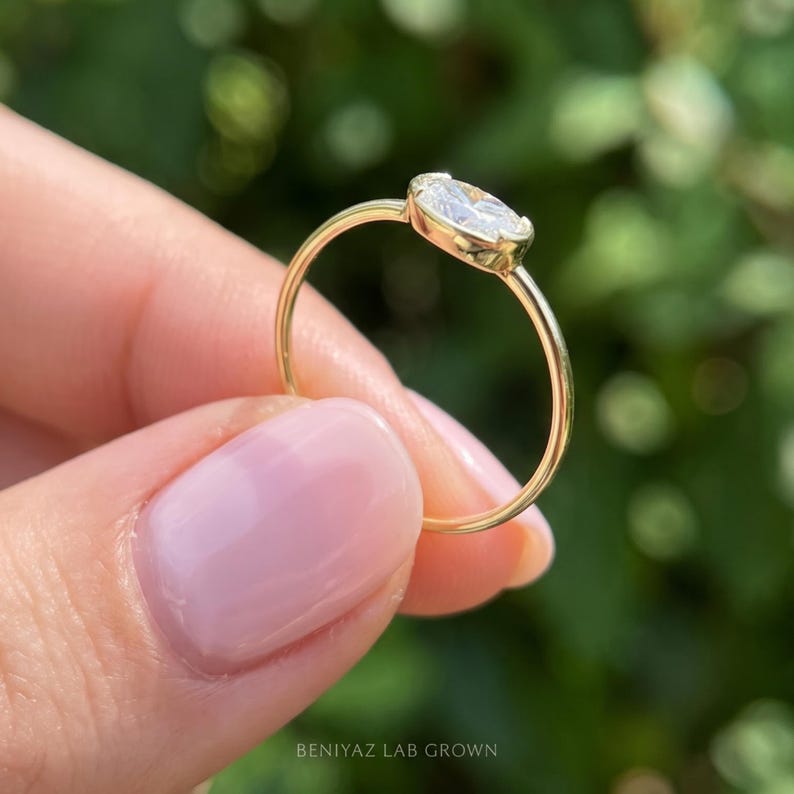 Angled view of a dainty oval bezel engagement ring held between fingertips. The horizontally positioned oval diamond sits in a polished gold setting above a slender band, with soft natural greenery in the background.