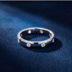 May include: A silver-toned metal band ring featuring multiple round, clear, bezel-set stones embedded along the outer surface. The text BENIYAZ LAB GROWN is engraved on the inner band. The ring has a polished, reflective finish and a smooth, circular shape.