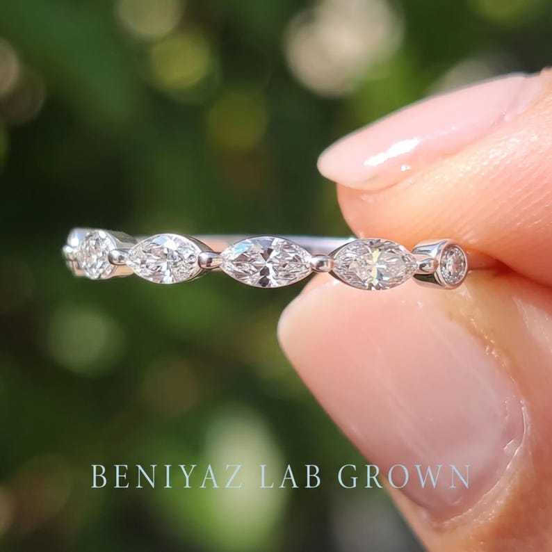 Macro front view of a delicate marquise diamond band held between fingertips. Three brilliant marquise cut lab grown diamonds with round accent stones form an elegant five-stone design on a slim polished metal ring.