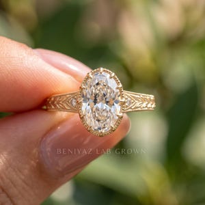 An oval diamond engagement ring photographed at a slight angle between fingertips. The image highlights the oval center stone, intricate side detailing and warm gold tone metal in natural daylight.