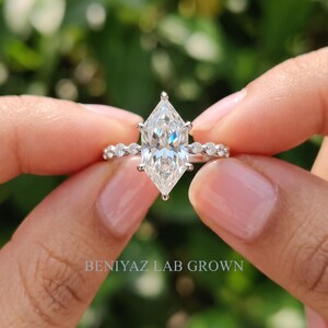 Detailed macro view of a Dutch marquise diamond engagement ring held between fingertips, showing the brilliant center diamond, six prong setting and symmetrical diamond accent design.