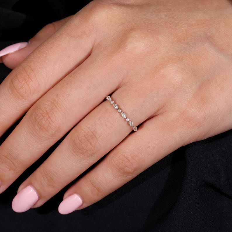 Close-up lifestyle photograph of a delicate lab grown diamond stacking ring worn on a woman's finger. The polished slim band features alternating marquise and round diamond accents arranged across the front. Soft natural skin tones, pale pink nails and understated styling highlight the fine details of this elegant half eternity wedding band.