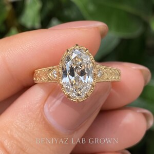 Macro front view of an oval diamond engagement ring held between fingertips. The sparkling oval center gemstone, smaller diamond accents and detailed band are shown against a soft green background.