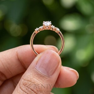 K&ouml;nnte beinhalten: Ein Ring aus Ros&eacute;gold mit einem zentralen, rund geschliffenen wei&szlig;en Stein in einer Krappenfassung, flankiert von zwei kleineren Steinen auf jeder Seite der Ringschiene. Der Metallring hat eine polierte Oberfl&auml;che und ein abgerundetes Profil.