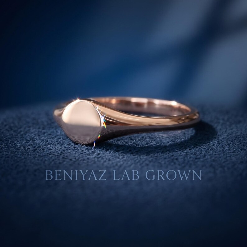 Side profile of plain round signet ring on blue background showing thin tapered band and rose gold tone high polish finish
