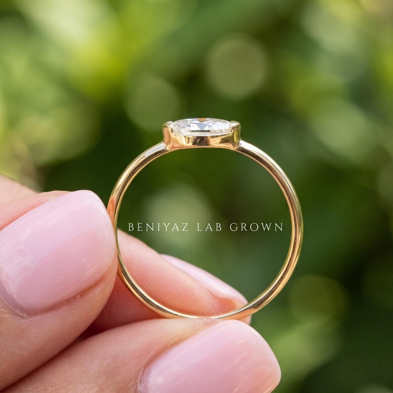 Close side view of an east west oval diamond engagement ring held between fingers outdoors. The polished gold band curves beneath the horizontally set oval gemstone, revealing the delicate bezel profile and clean minimalist construction.