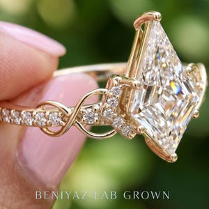 Extreme side-angle close-up of a geometric kite diamond engagement ring, highlighting the elevated center stone, prong structure and interwoven pavé diamond details. The gold tone band and delicate setting are sharply focused against a soft garden background.