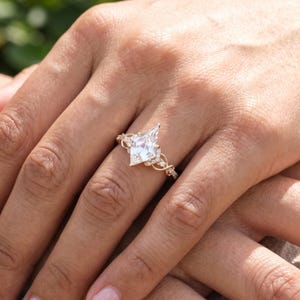 Close-up of a woman's hand wearing a kite shaped lab grown diamond engagement ring against soft neutral fabric. The distinctive pointed center diamond stands out above the sparkling pavé band, showing the ring's elegant scale on the finger.