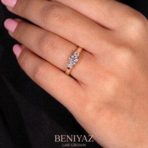 May include: A gold ring featuring three round-cut clear stones set in a row. The centre stone is larger than the two flanking stones. The text BENIYAZ LAB GROWN is printed in white at the bottom of the image against a black background.