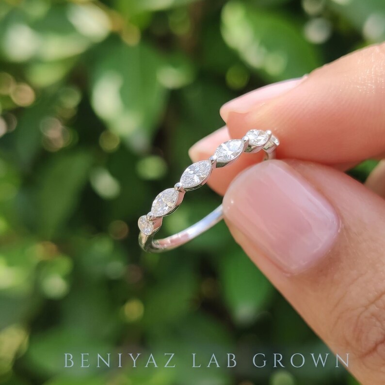 Three-quarter side view of a marquise and round diamond stacking band held between fingers. The angled photograph reveals the diamond prong settings, slim curved profile and polished white metal shank against a soft green background.