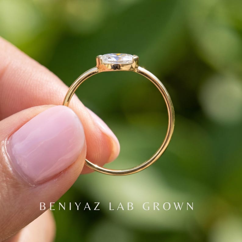 Full side profile of an east west oval diamond ring held upright between fingers. The photograph shows the thin circular gold band, low raised bezel setting and horizontally oriented center stone against a blurred outdoor background.
