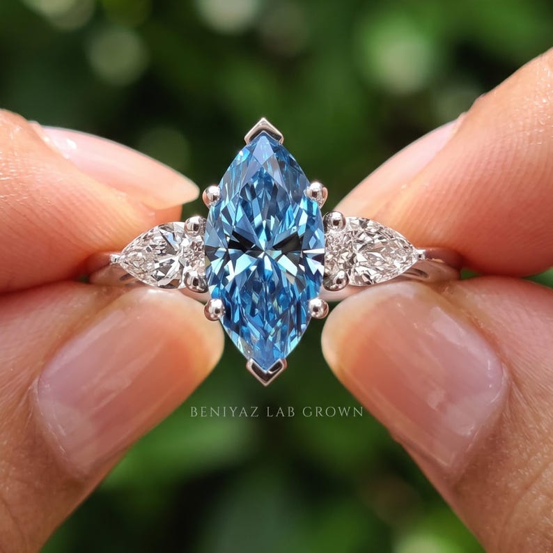 Close front hand held view of a blue marquise lab grown diamond ring highlighting the elongated blue center and white pear side diamonds.