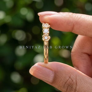 May include: A gold ring featuring a band with two flower-shaped settings, each containing a small, clear, round-cut stone. The text BENIYAZ LAB GROWN is visible across the center of the image. The ring has a polished, reflective metallic finish.