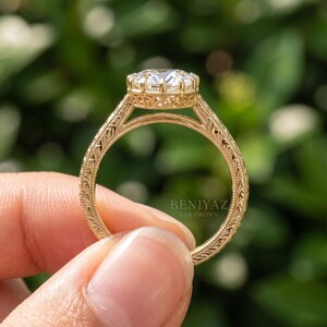 Full side view of an oval diamond engagement ring held vertically between fingertips. The raised center stone setting and decorative diamond accents along the yellow gold tone shank are visible.