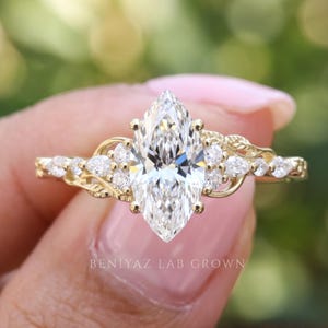 Macro view of a unique marquise diamond ring featuring a prominent pointed center stone and sparkling leaf cluster accents. The polished gold band and delicate gemstone arrangement are photographed between fingertips against a naturally blurred outdoor background.