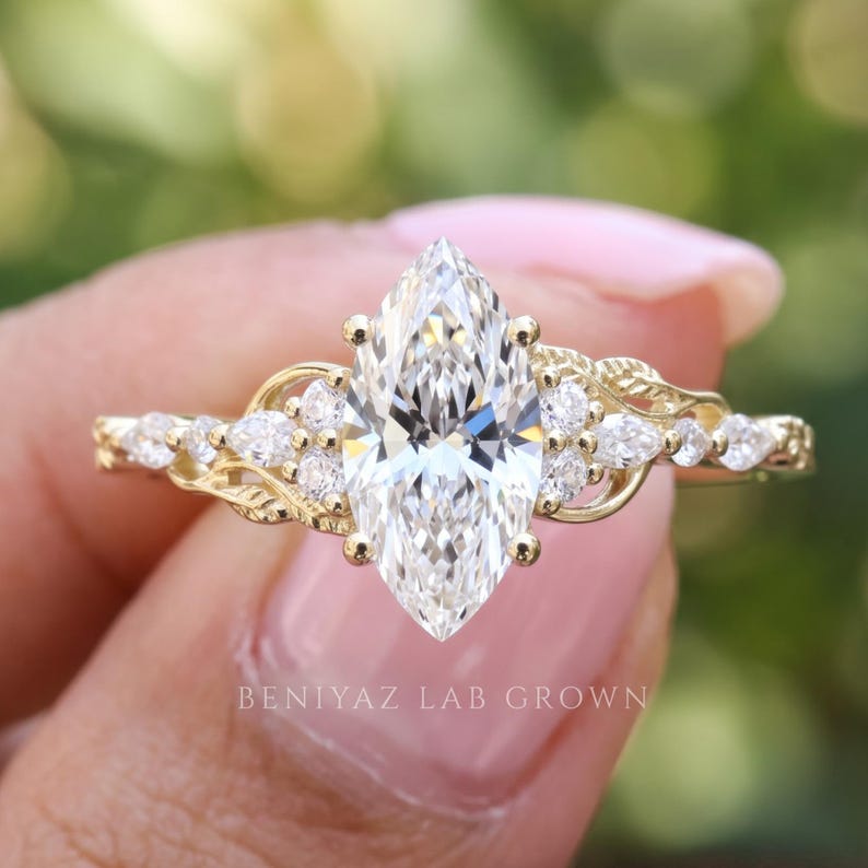 Macro view of a unique marquise diamond ring featuring a prominent pointed center stone and sparkling leaf cluster accents. The polished gold band and delicate gemstone arrangement are photographed between fingertips against a naturally blurred outdoor background.