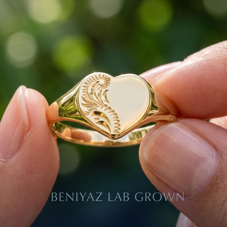 Close up heart signet ring held by hand, showing the smooth polished face, floral scroll detail and tapered yellow gold tone shank
