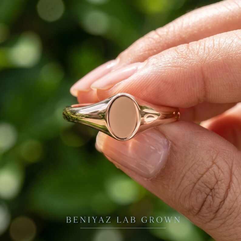 Close up oval signet ring held between fingers showing blank high polish face, tapered shoulders and delicate band
