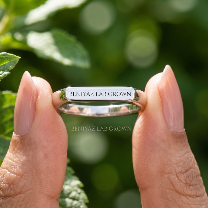 Hand holding a custom engraved wedding band with personalized name and date detail, minimalist polished ring photographed against a natural green background