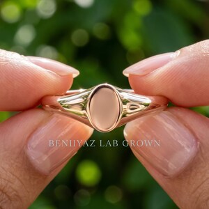 Petite plain oval signet ring held between fingers with smooth polished face and minimal tapered band design