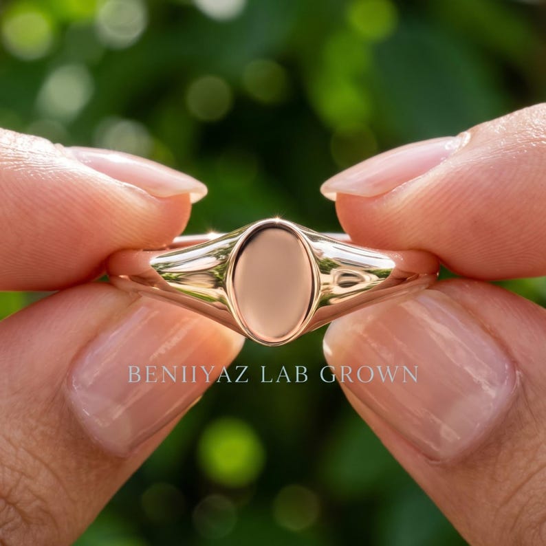 Petite plain oval signet ring held between fingers with smooth polished face and minimal tapered band design