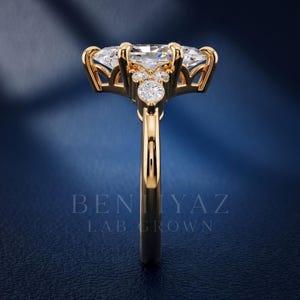 May include: A gold ring featuring a large marquise-cut center stone held by prongs, accented by smaller round-cut stones along the gallery and band. The text BENIYAZ LAB GROWN is visible in the lower portion of the image, centered below the ring.