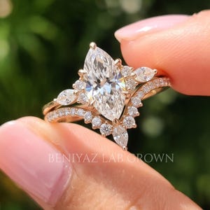 Detailed macro view of a marquise diamond engagement ring set held between fingertips. The elongated pointed center gemstone, sparkling vintage inspired side accents and matching curved wedding band create an intricate bridal design.