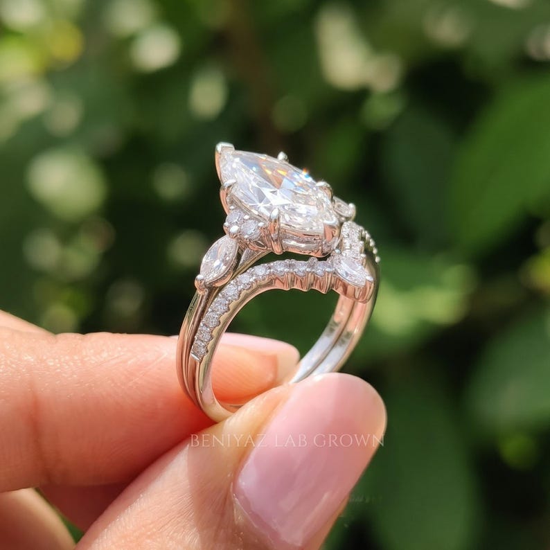 Side-angle photograph of a marquise lab grown diamond engagement ring set held upright between fingertips. The raised center stone, decorative prong setting and curved diamond wedding band show how the two rings fit together.