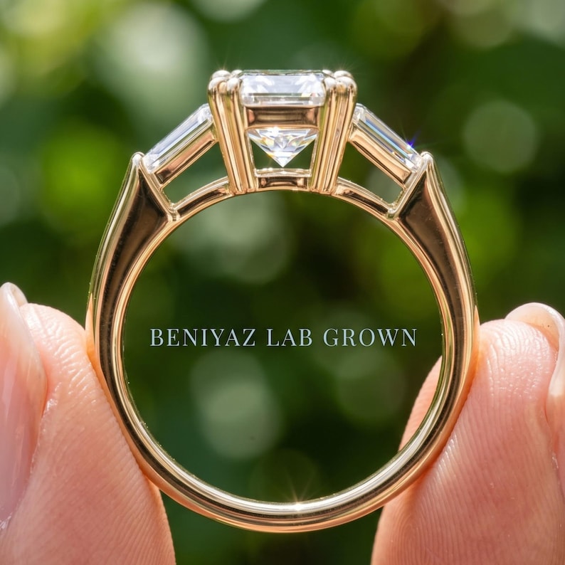 Rear view of emerald cut three stone lab grown diamond ring held by hand, showing raised open gallery, tapered side stones and polished gold shank
