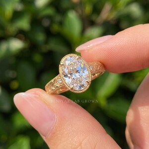 Three-quarter close-up of an oval diamond engagement ring held between fingertips. The photograph highlights the oval center stone, sparkling side diamonds and intricate yellow gold tone band in natural outdoor light.
