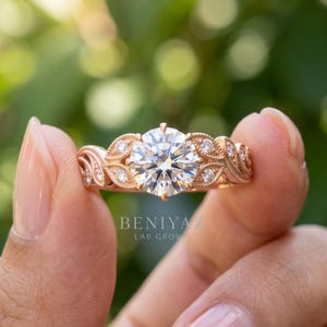 Hand displaying a round lab grown diamond engagement ring in yellow gold with a classic multi stone design and bright outdoor green backdrop