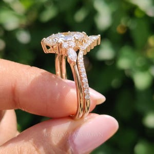 Full side profile of a marquise diamond bridal set held vertically between fingertips outdoors. The photograph highlights the elevated center setting, open gallery and two slender rose gold tone bands with delicate diamond accents.