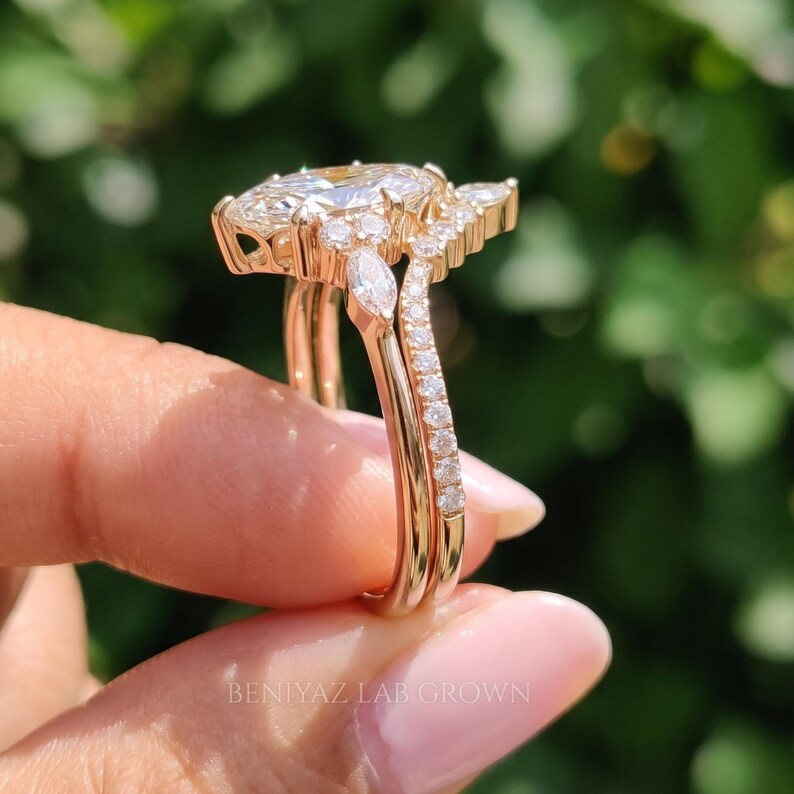 Full side profile of a marquise diamond bridal set held vertically between fingertips outdoors. The photograph highlights the elevated center setting, open gallery and two slender rose gold tone bands with delicate diamond accents.