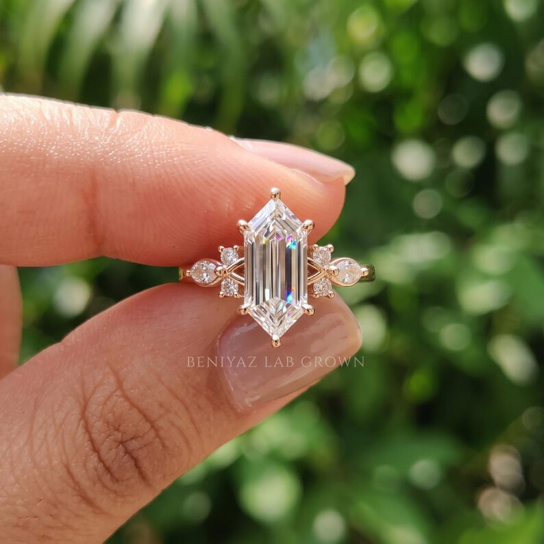 Elongated hexagon engagement ring held between fingers with step cut lab grown diamond center and decorative marquise round sides