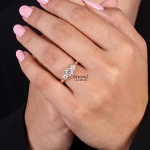 May include: A gold ring featuring three round-cut clear stones in a prong setting. The text BENIYAZ LAB GROWN is printed in black near the ring. The band is thin and polished gold, showcasing a classic three-stone design with a central larger stone.