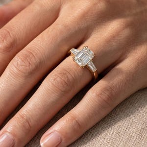 Woman wearing an emerald cut lab grown diamond engagement ring with three stone setting and tapered diamond side stones on yellow gold band