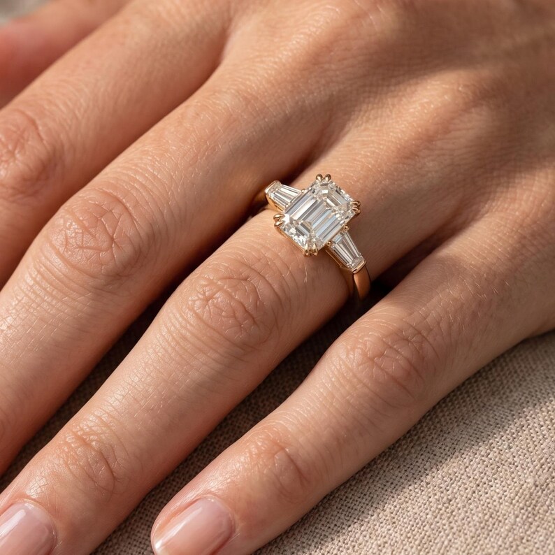 Woman wearing an emerald cut lab grown diamond engagement ring with three stone setting and tapered diamond side stones on yellow gold band