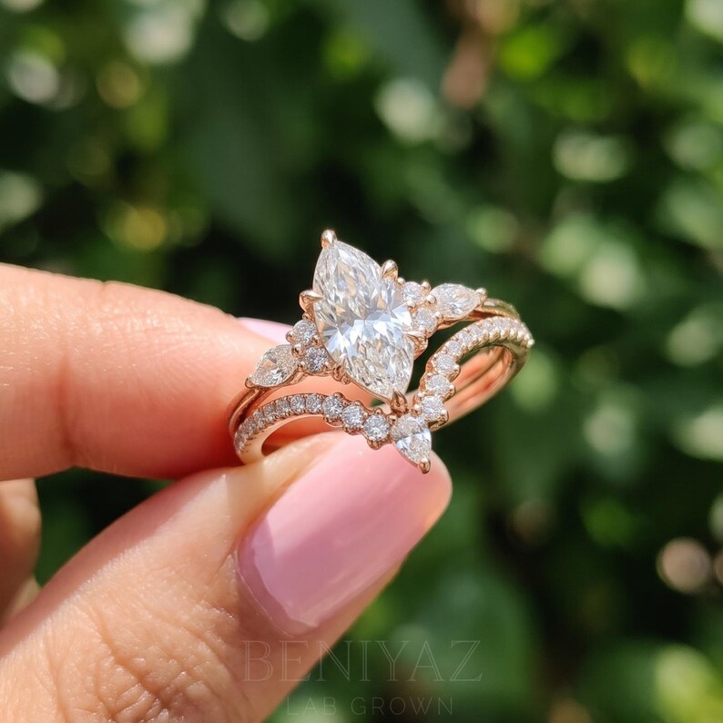 Side view of a marquise engagement ring set held between fingertips outdoors. The elevated diamond setting and two closely fitted rose gold tone bands feature sparkling accent stones along their upper sections.