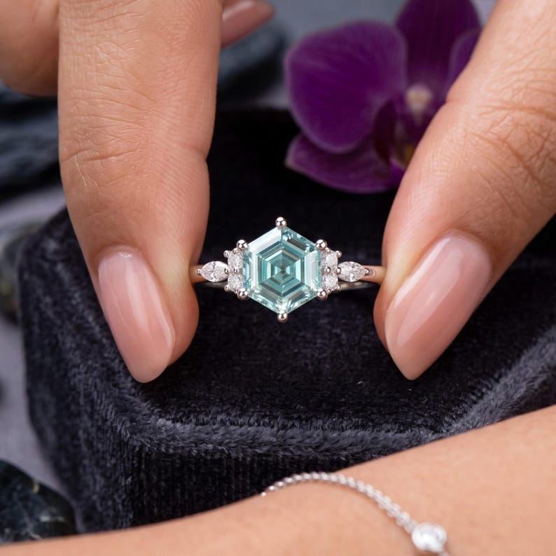 Cluster ring for women shown on hand with blue hexagon moissanite center, sparkling side stones and modern geometric design.