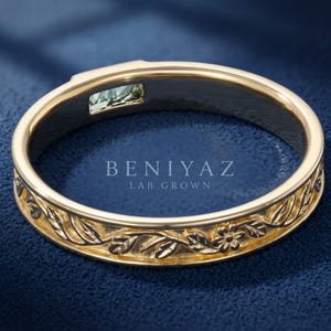 Puede incluir: Un anillo de oro con un patr&oacute;n floral y de hojas grabado en la superficie exterior. El interior de la banda contiene una peque&ntilde;a piedra preciosa rectangular de color verde claro y el texto BENIYAZ LAB GROWN grabado en el centro.