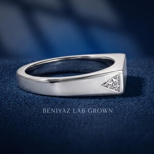 Puede incluir: Un anillo de metal plateado con una banda plana y ancha que presenta una piedra transparente de corte triangular incrustada a ras de la superficie. El texto BENIYAZ LAB GROWN aparece debajo del anillo, que tiene un acabado pulido y brillante.