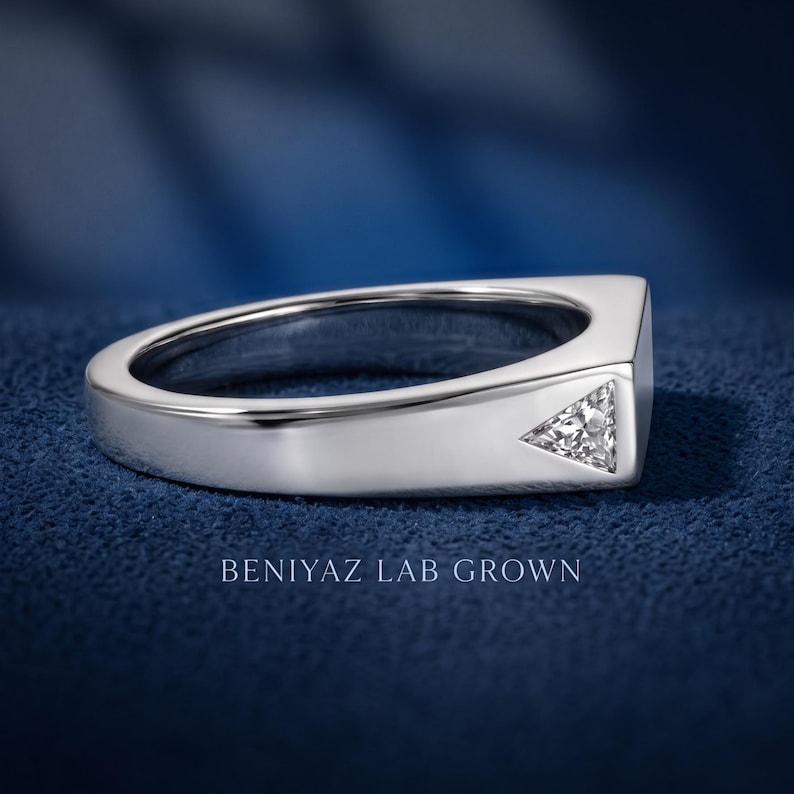 Puede incluir: Un anillo de metal plateado con una banda plana y ancha que presenta una piedra transparente de corte triangular incrustada a ras de la superficie. El texto BENIYAZ LAB GROWN aparece debajo del anillo, que tiene un acabado pulido y brillante.