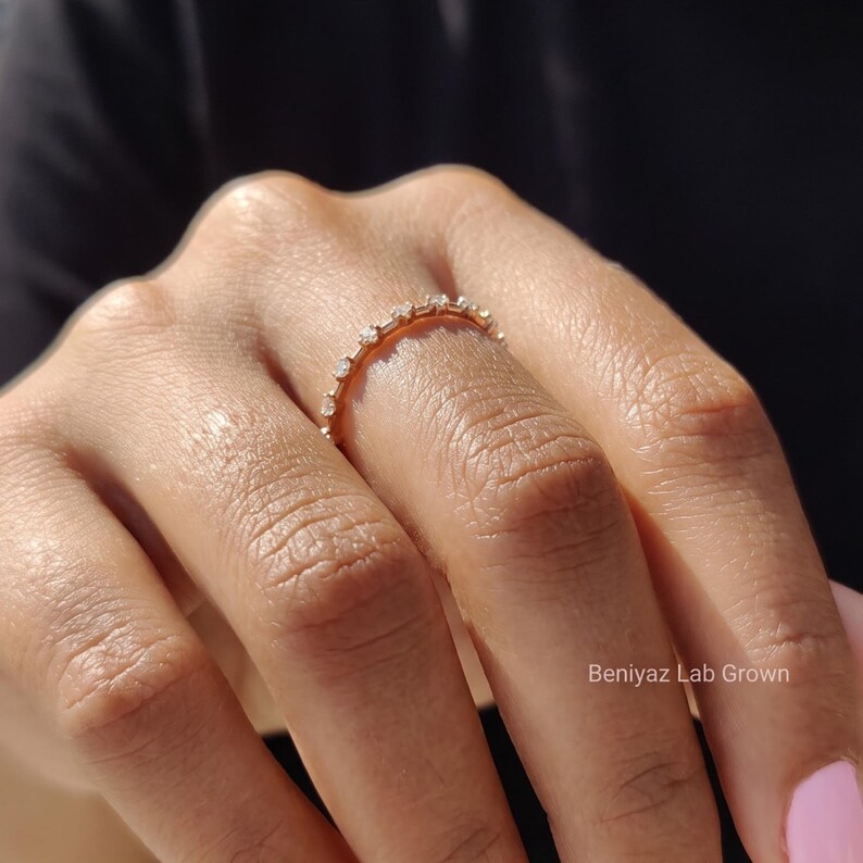 Close-up of a woman's hand wearing a slim marquise moissanite wedding band. The delicate ring adds subtle sparkle to a minimalist look, styled with a dark sleeve and natural pink nails.